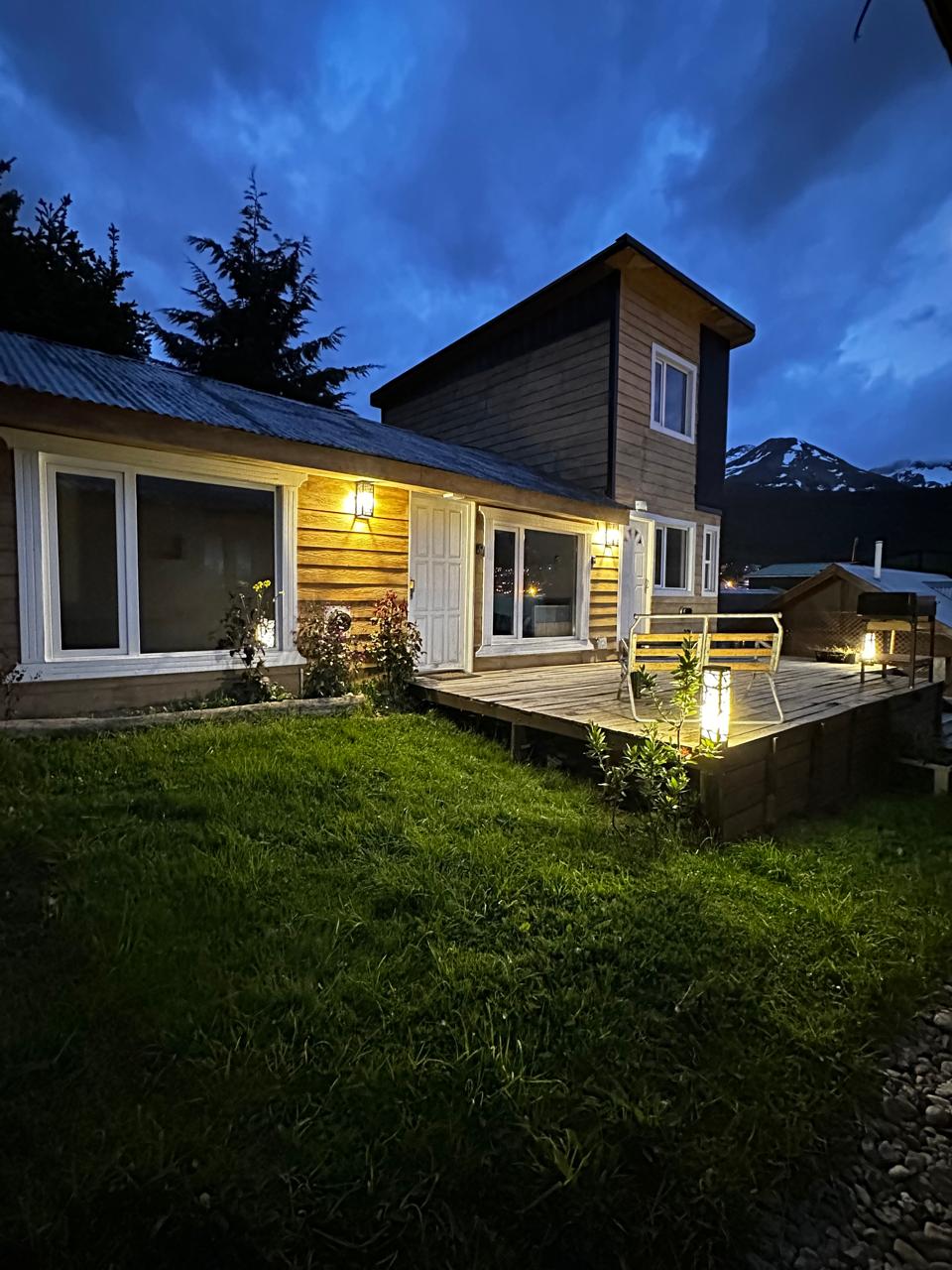The exterior of a wooden house is shown during twilight, featuring large windows and a well-lit deck. Surrounded by greenery, the area is complemented by subtle outdoor lighting. A mountain range is faintly visible in the background under a cloudy sky.