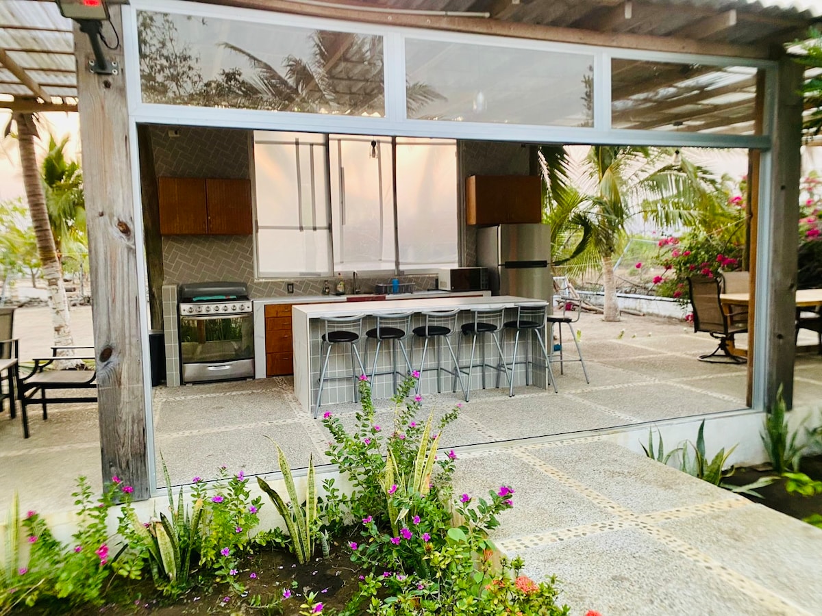 An outdoor kitchen area features a spacious countertop with bar stools, complemented by wooden cabinets and modern appliances. Lush greenery with flowering plants surrounds the space, creating a natural atmosphere. Large windows provide ample lighting and an open view to the surroundings.