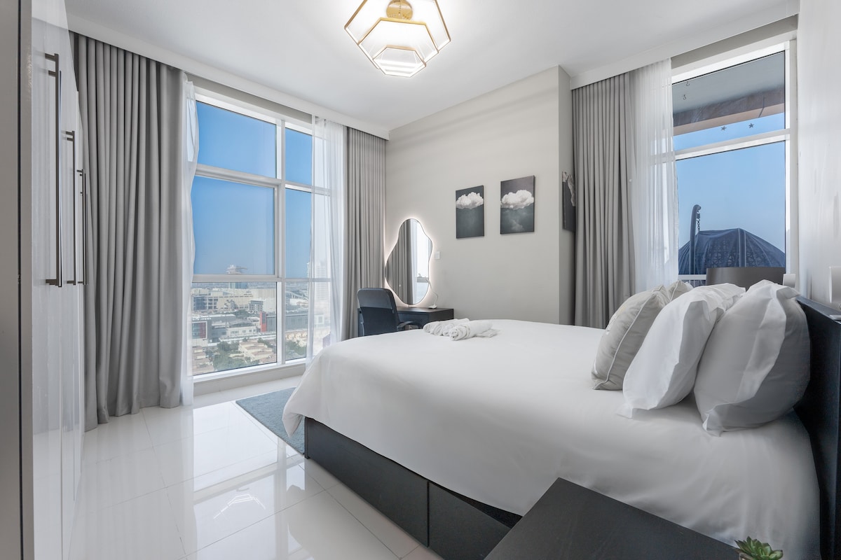 The bedroom features a king bed with fresh linens and multiple pillows. Large windows offer natural light and city views. A mirror and a desk are positioned against the wall, with soft curtains framing the windows, creating a serene environment.