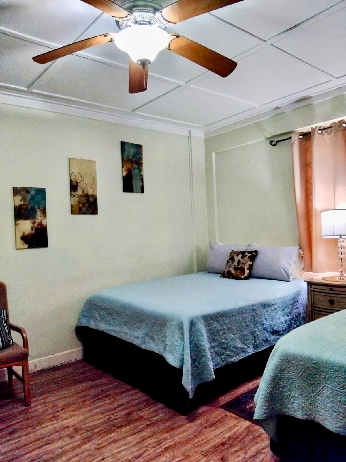 A cozy room features two queen-size beds covered with light blue bedspreads. A bedside lamp and a side table are positioned next to one bed. Wall art decorates the space, and a chair is placed in the corner. Natural light filters through the window with soft curtains.