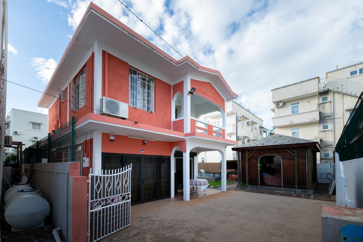 A spacious two-story bungalow is shown, featuring a vibrant exterior in soft red tones. An outdoor area includes a paved driveway and a kiosk with a sloped roof. Clear skies and scattered clouds create a serene setting, with air conditioning units visible on the façade.