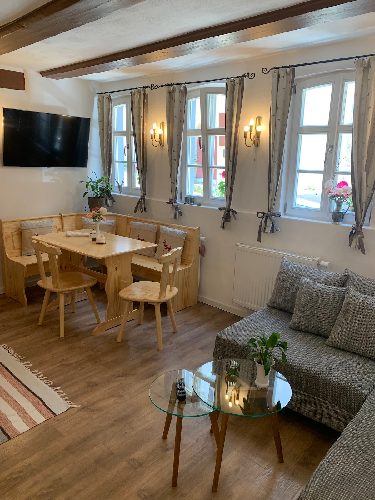 The living area features a cozy seating arrangement with a grey sofa and a glass coffee table. A wooden dining table surrounded by four chairs is positioned near bright windows adorned with sheer curtains. Small potted plants add a touch of greenery.