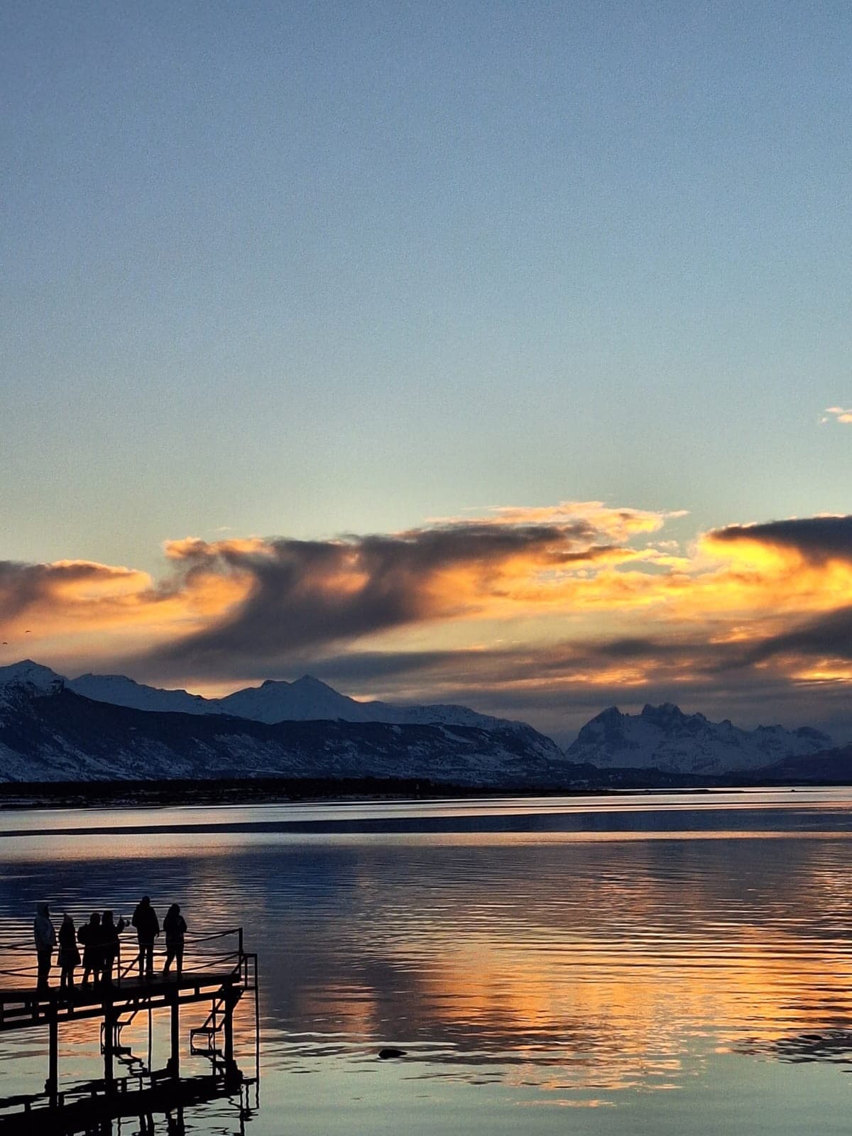 A serene waterscape is depicted, featuring calm waters reflecting warm sunset hues. Silhouetted figures stand on a wooden pier, overlooking distant mountains under a colorful sky. The scene captures the tranquility of nature, inviting relaxation and contemplation.