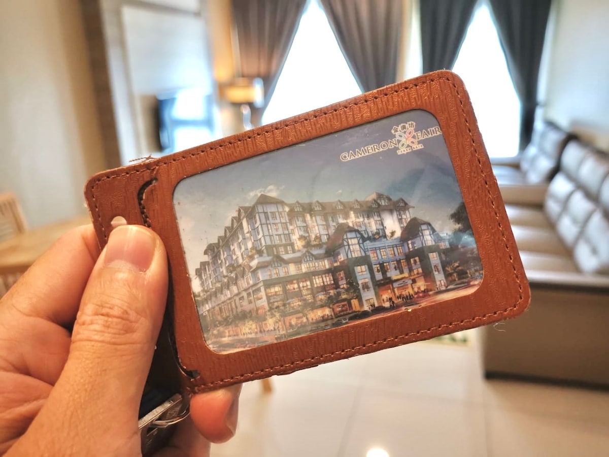 A hand is holding a key card in a brown leather case that displays an image of the Cameron Fair complex. The background features an elegantly furnished living area with neutral-colored furniture and large windows allowing natural light to enter.