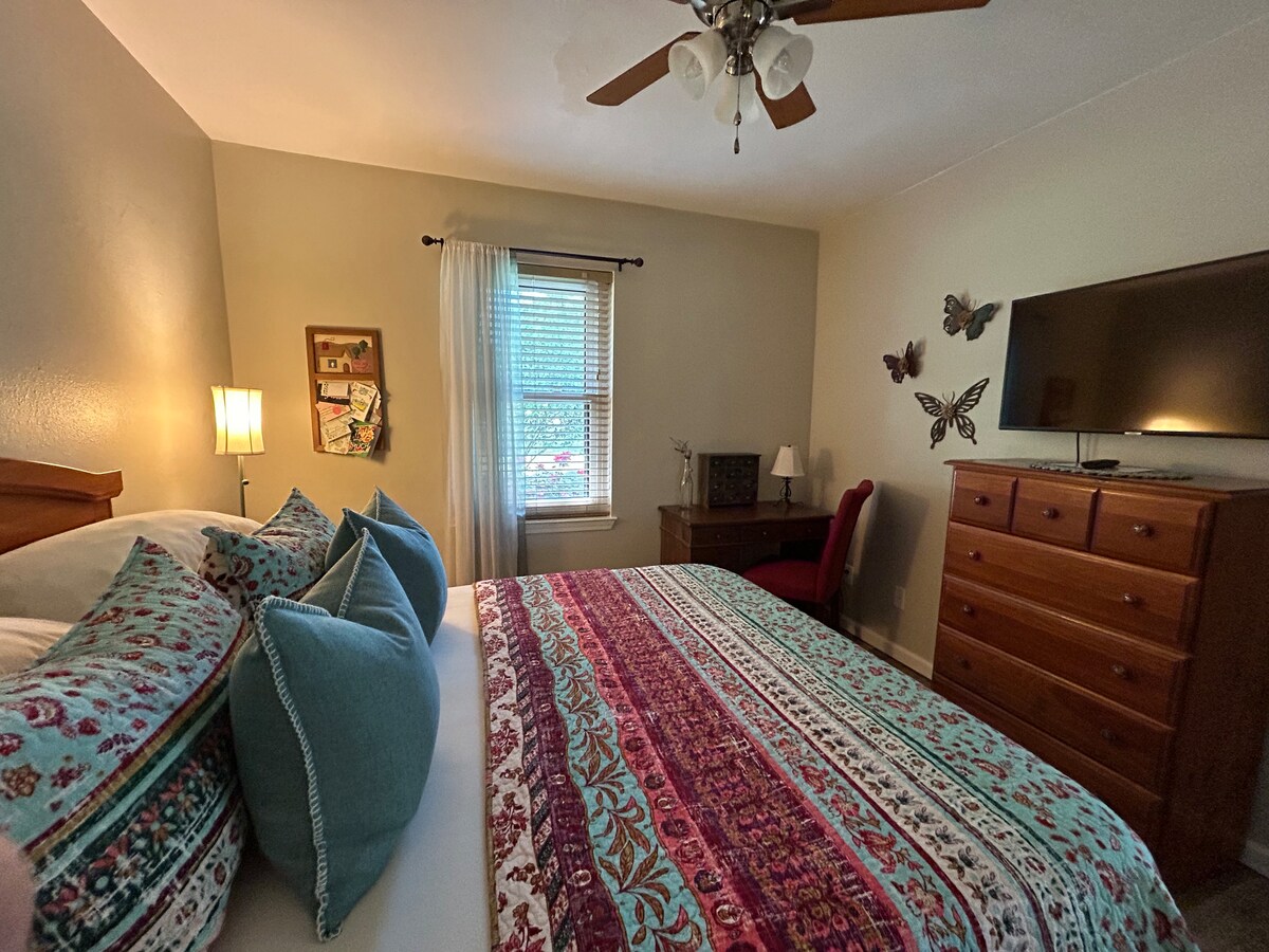 A cozy bedroom features a spacious bed adorned with a colorful patterned throw and decorative pillows. Natural light enters through a window with sheer drapes. A wooden dresser and desk are included, accompanied by butterfly wall art for added charm.