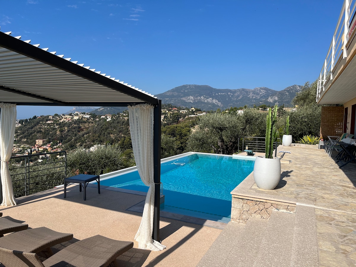 An inviting outdoor area features a private swimming pool surrounded by a paved terrace. A shaded pergola offers a comfortable seating space with loungers. Lush greenery and mountain views enhance the serene setting, with a clear blue sky overhead.