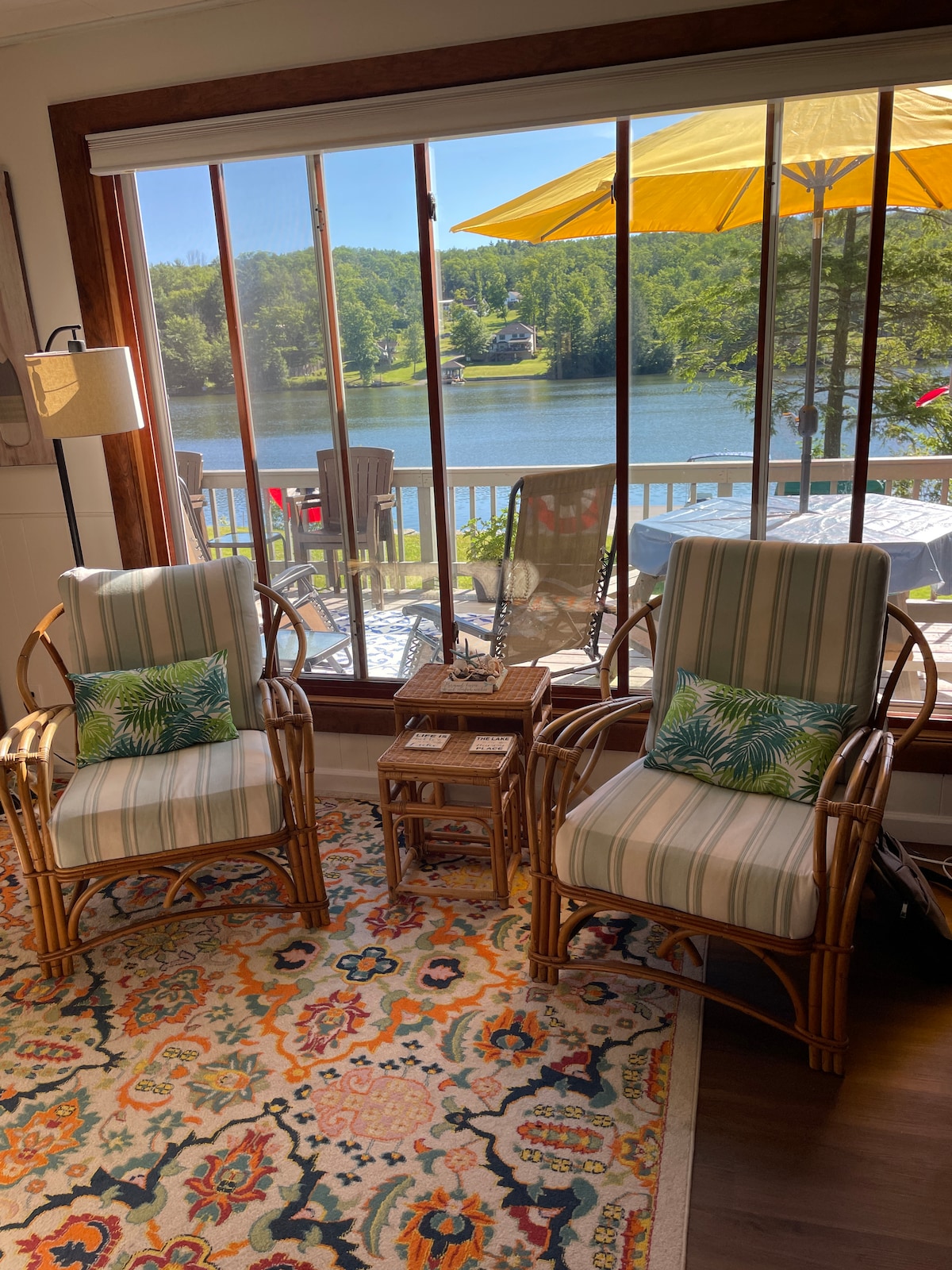 A bright sitting area features two striped chairs adorned with patterned cushions. A small table rests between them, surrounded by a colorful area rug. Large windows provide a view of the lake, with an outdoor dining space visible. A yellow umbrella casts shade on the patio.