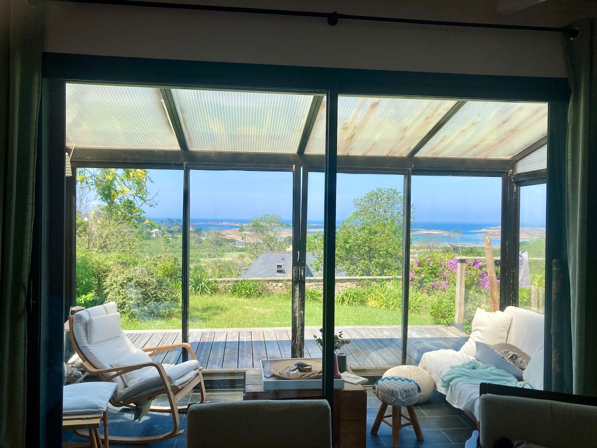 A spacious and bright veranda features large sliding glass doors, offering views of the landscape and ocean. Comfortable seating, including a rocking chair, is arranged thoughtfully, inviting relaxation. Natural light floods the space, highlighting the greenery outside and the tranquil atmosphere.