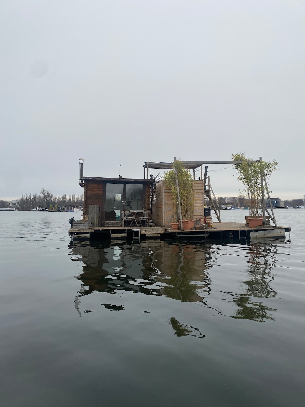 A floating houseboat is anchored on calm waters, featuring a wooden exterior with large windows and a small deck area. Potted plants are placed on the deck, adding a touch of greenery. The overcast sky reflects in the water, creating a serene atmosphere.