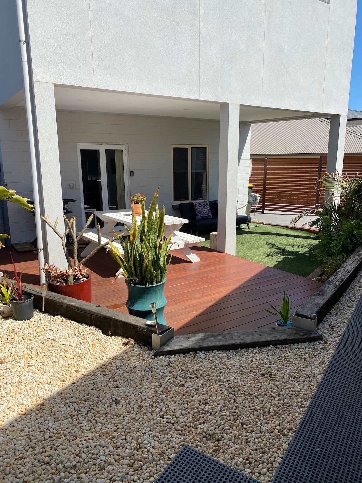 A secure back yard is featured, showcasing a timber deck with an outdoor seating area. Green tropical plants add a natural element, while decorative gravel complements the space. French doors lead to the apartment, enhancing the connection between indoors and outdoors.