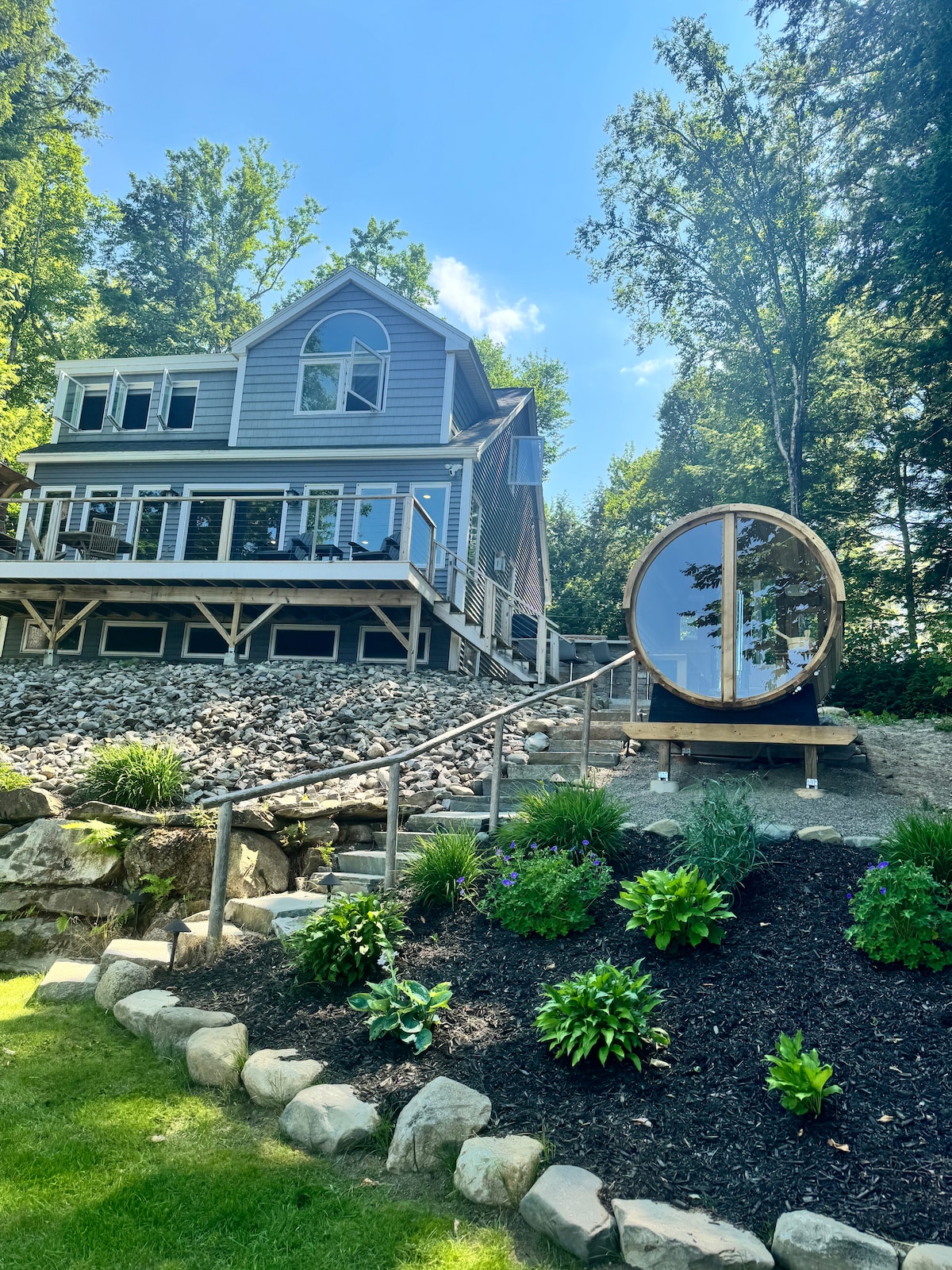 The exterior of a rustic/modern lake house is presented, featuring a large deck that overlooks the water. A circular sauna pod is situated in the foreground, framed by landscaped gardens with a variety of plants. The surrounding trees provide a natural backdrop to the home.