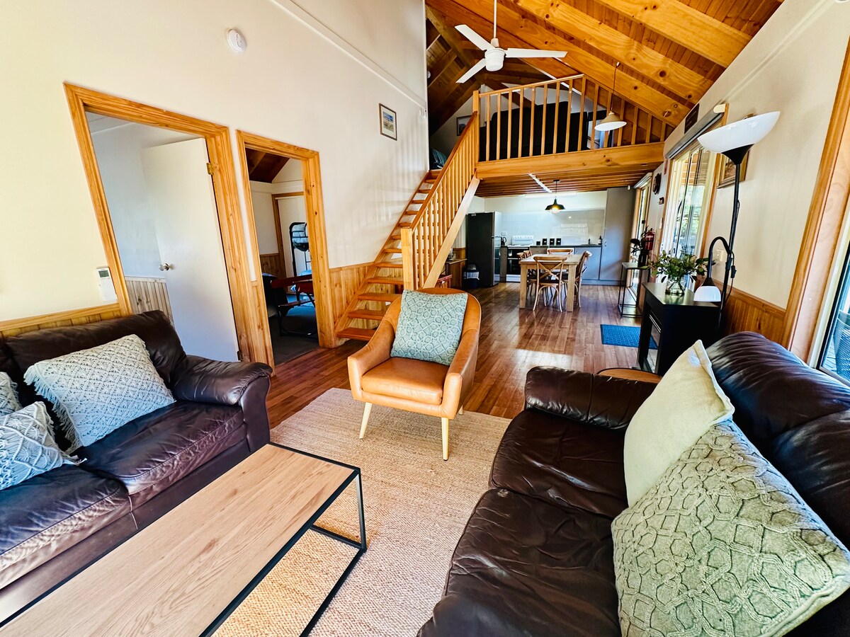 A spacious living area showcases dark leather sofas with decorative pillows, paired with a light coffee table. Natural light streams through large windows, illuminating the wooden ceiling and the open plan leading to a dining area with a staircase in the background.