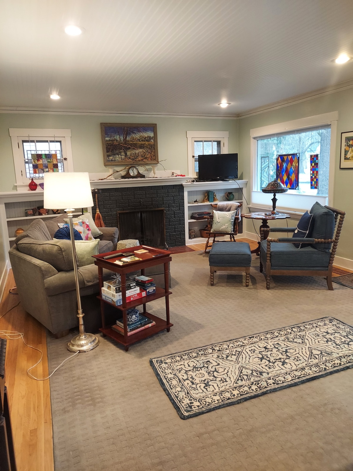 A cozy living room is presented, featuring a gray sofa and two blue armchairs positioned around a small table. A decorative fireplace and a television are visible on the wall, while stained glass accents add color. A soft area rug lies beneath, enhancing the inviting space.