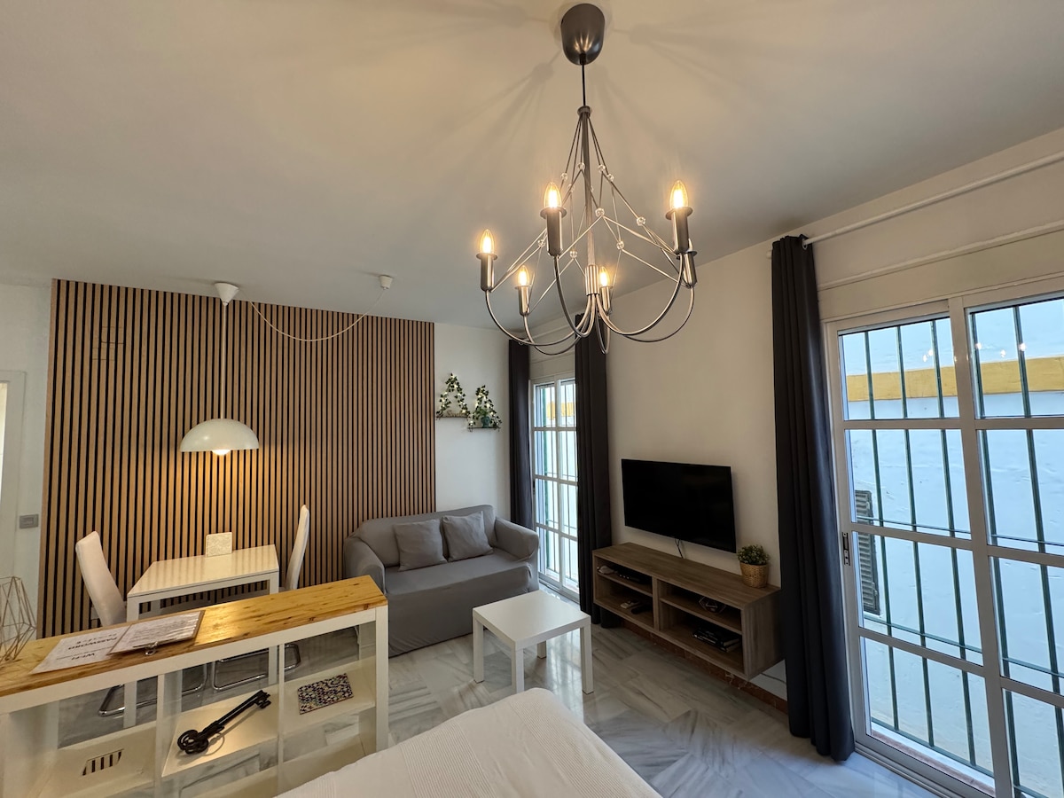 A functional living space features a stylish chandelier and wooden accent wall. A dining table with chairs is paired with a gray sofa, while a television is mounted on the wall near a light-colored coffee table. Large windows provide natural light and outdoor views.