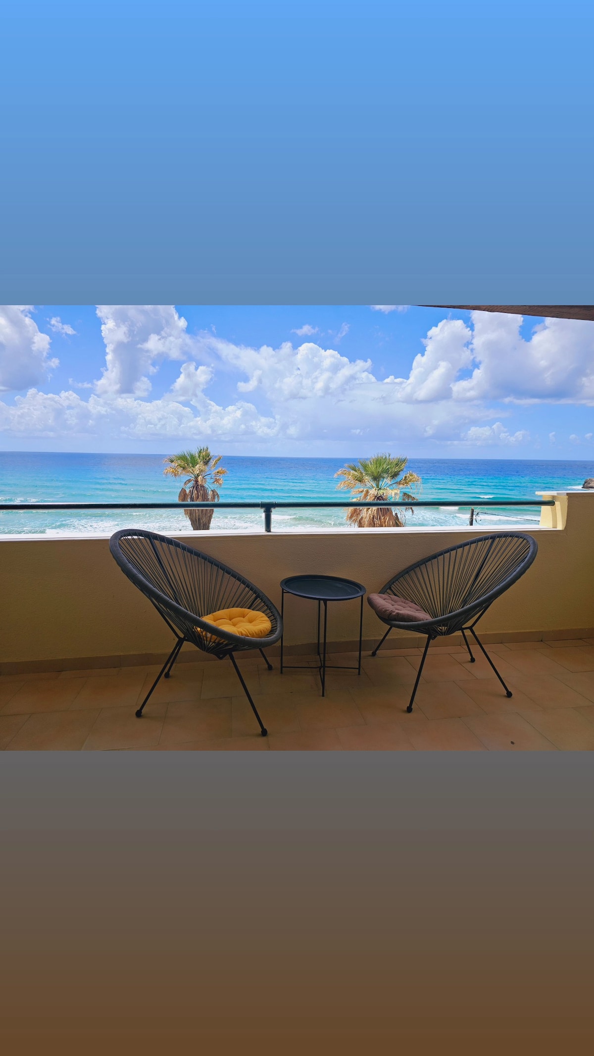 A balcony area features two modern chairs alongside a small table. The view showcases the expansive sea and clear skies, with palm trees visible in the background, creating a serene setting for relaxation.