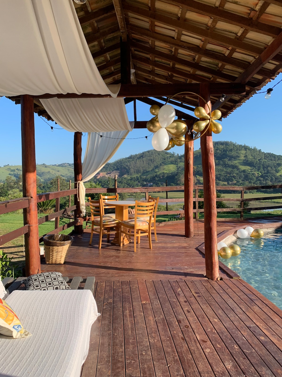 A wooden deck area features a dining table with four chairs, surrounded by a scenic view of the mountains. Soft drapes flutter gently in the breeze, while nearby, a cozy seating arrangement invites relaxation beside the shimmering pool.