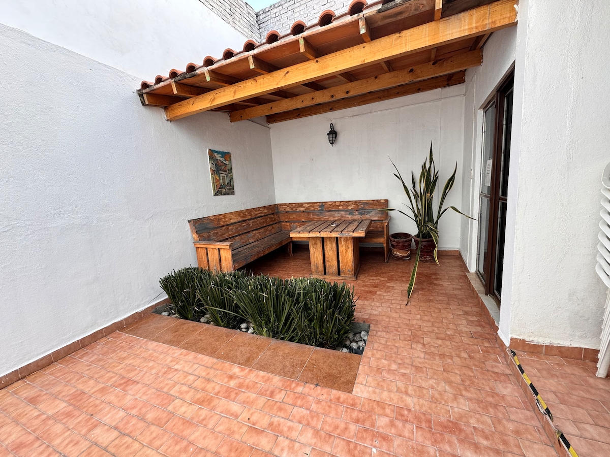 An open patio area features wooden seating around a rectangular table, with a low planter filled with greenery at the center. The terracotta-tiled floor and white walls create a clean and spacious environment, complemented by natural light from the adjacent doorway.