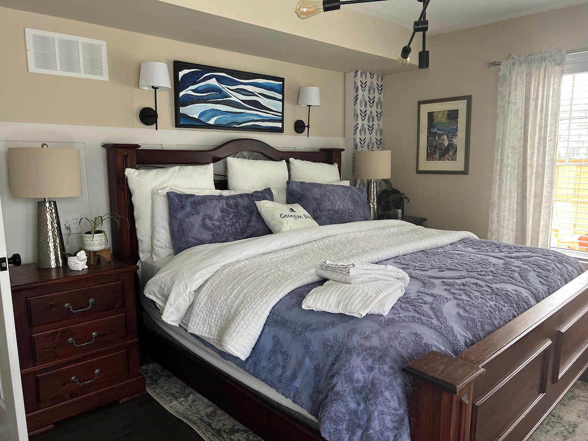 A spacious bed is layered with soft, textured bedding in calming shades of blue and white. Two bedside lamps provide gentle illumination, and artwork depicting waves adorns the wall above. Large windows allow natural light to brighten the room, enhancing its inviting ambiance.