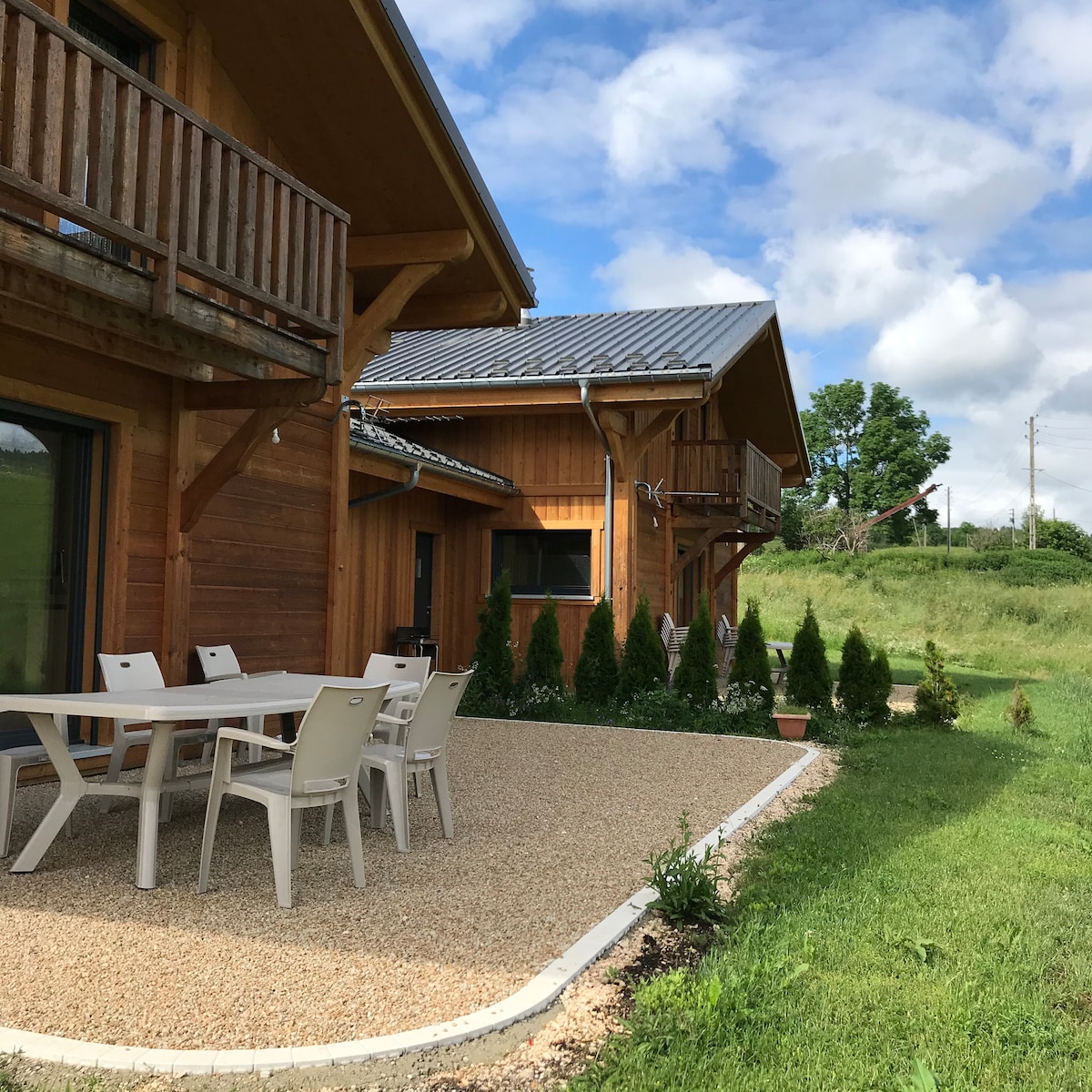 An outdoor terrace features a table with six chairs, surrounded by neatly arranged gravel and green landscaping. The wooden structure of the chalet blends harmoniously with the natural surroundings, while planters and low shrubs offer a touch of greenery along the edge.