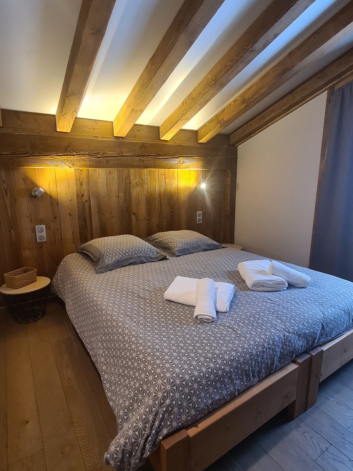 A cozy bedroom features a spacious bed with patterned bedding. Soft lighting from wall fixtures creates a warm ambiance. Fresh towels are neatly arranged at the foot of the bed, complementing the rustic wooden wall and beams visible in the ceiling.