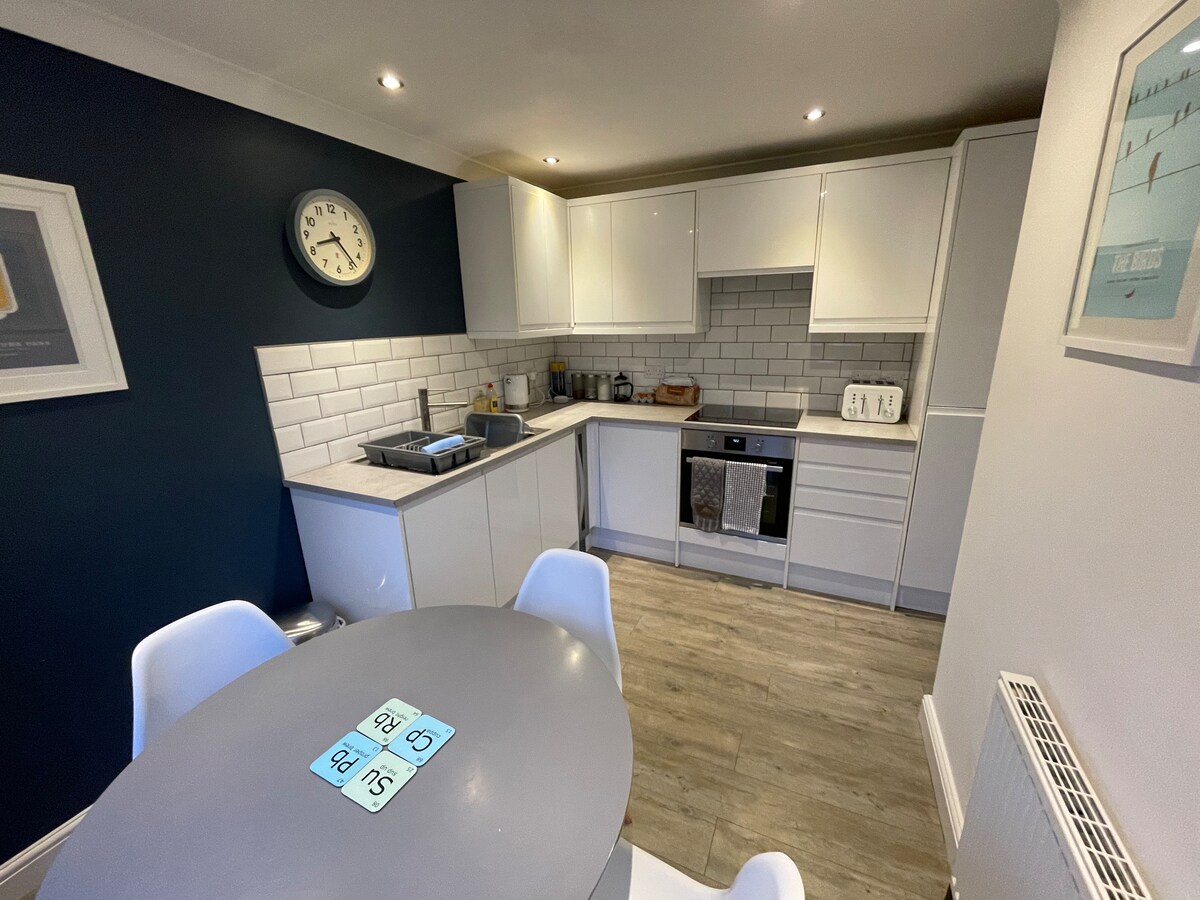 A modern kitchen features sleek white cabinetry contrasted by a deep blue accent wall. A circular dining table with light chairs is positioned centrally. Stainless steel appliances and a minimalist backsplash enhance the space, while warm lighting creates an inviting environment.