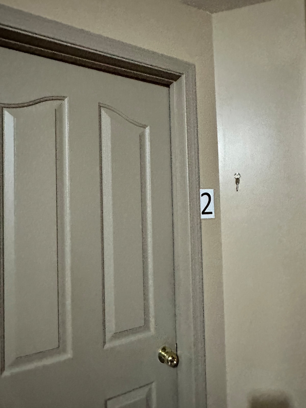 A light-colored door with a modern design is displayed prominently. A number '2' is affixed to the left side of the door, indicating the room number. A key hook is mounted on the wall beside the door, providing a convenient spot for hanging keys.