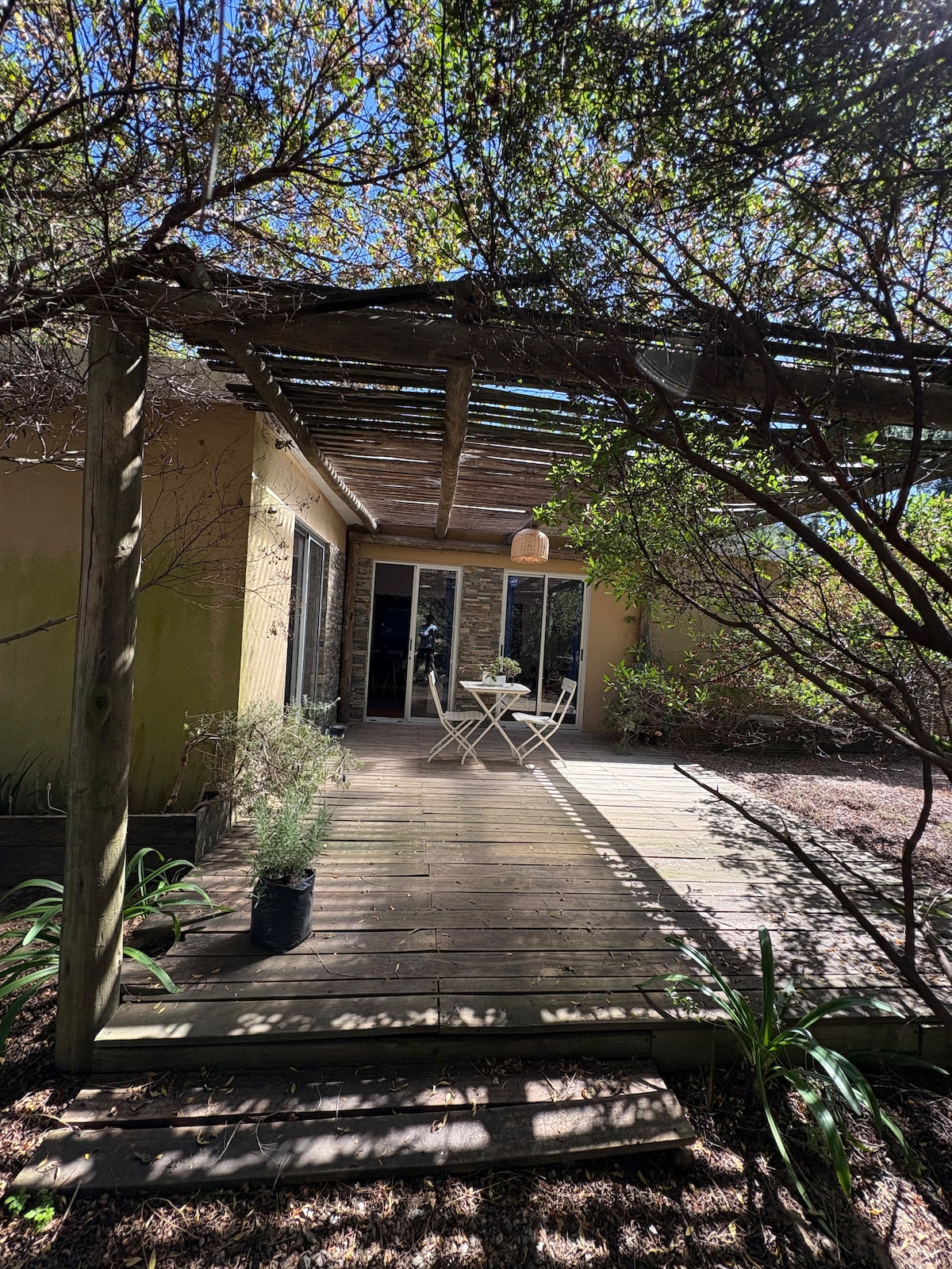 A wooden deck is framed by a pergola and surrounded by greenery, providing an inviting outdoor space. Two small chairs and a table are positioned for enjoyment, with natural light filtering through the overhanging foliage.