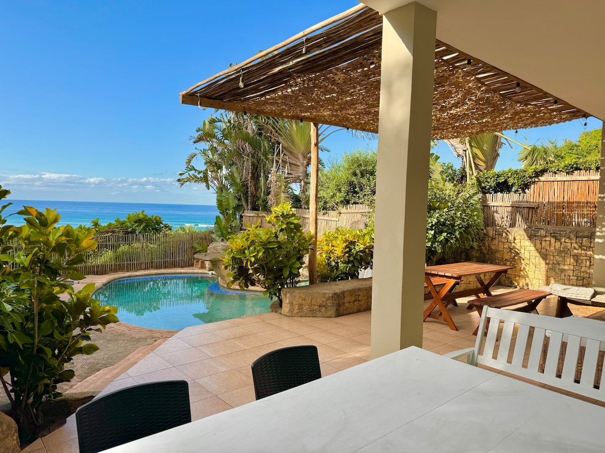 A shaded outdoor dining area is visible, featuring a white table and dark chairs. A lush garden surrounds a turquoise swimming pool, with ocean views beyond. Sunlight filters through palm leaves, creating a serene atmosphere for relaxation.