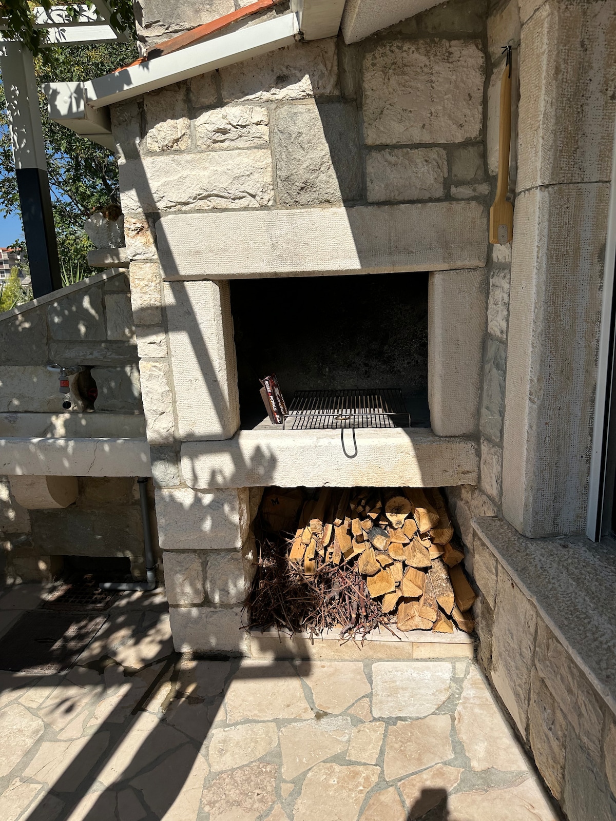 A traditional stone barbecue structure is depicted, featuring a built-in grill above a neatly stacked pile of firewood. Sunlight filters through nearby trees, casting dappled shadows on the surrounding stone patio.