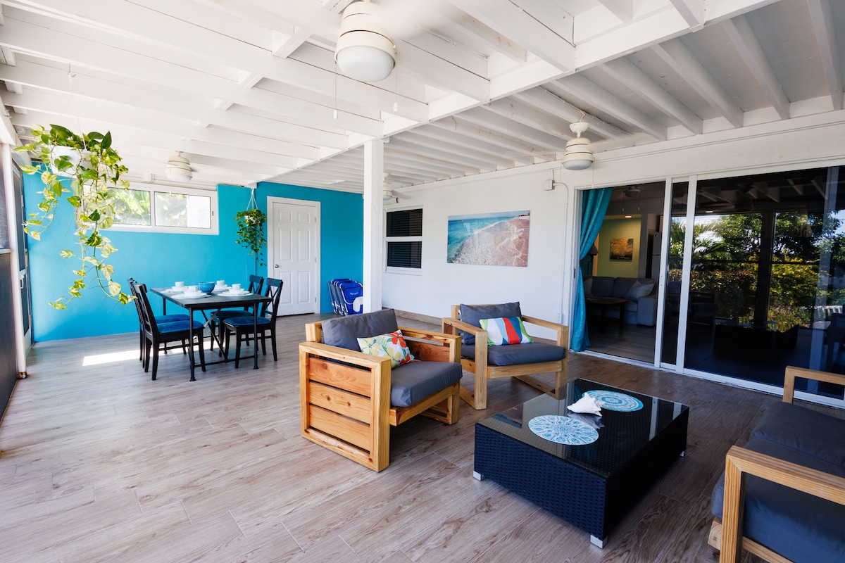 A spacious screened veranda is showcased, featuring comfortable seating arrangements with wooden furniture and colorful cushions. A dining area with a large table is visible, along with tropical plants and light-colored walls that enhance the bright atmosphere. Sliding glass doors lead to the living area.