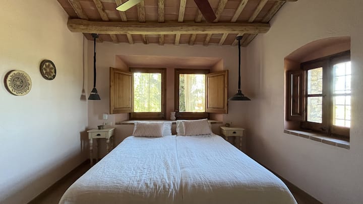 Detail of the bedroom with a double bed next to windows with wooden shutters that let in natural light. A warm and serene atmosphere beneath a ceiling with traditional beams.