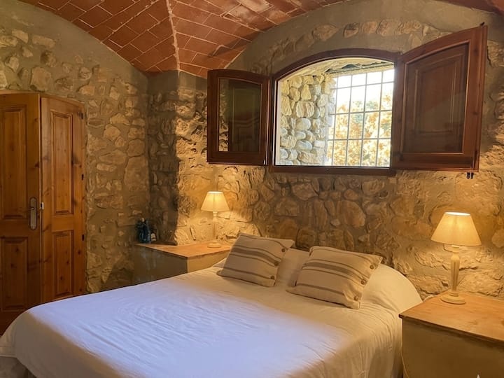 Ground-floor bedroom with stone walls and a vaulted brick ceiling, creating a warm and cosy atmosphere. Light streams through the window, framed by wooden shutters, creating a peaceful space for rest.