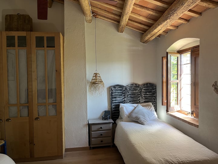 Detail of one of the single beds by the window, bathed in natural light, with simple furniture that maintains the rustic style of the house.