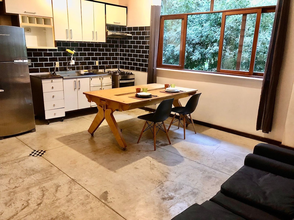 A bright kitchen area features a wooden dining table set for two, complemented by sleek black chairs. A contemporary kitchen with white cabinets and dark tiles is visible, along with large windows that open to a green view outside. The polished concrete floor adds a modern touch.
