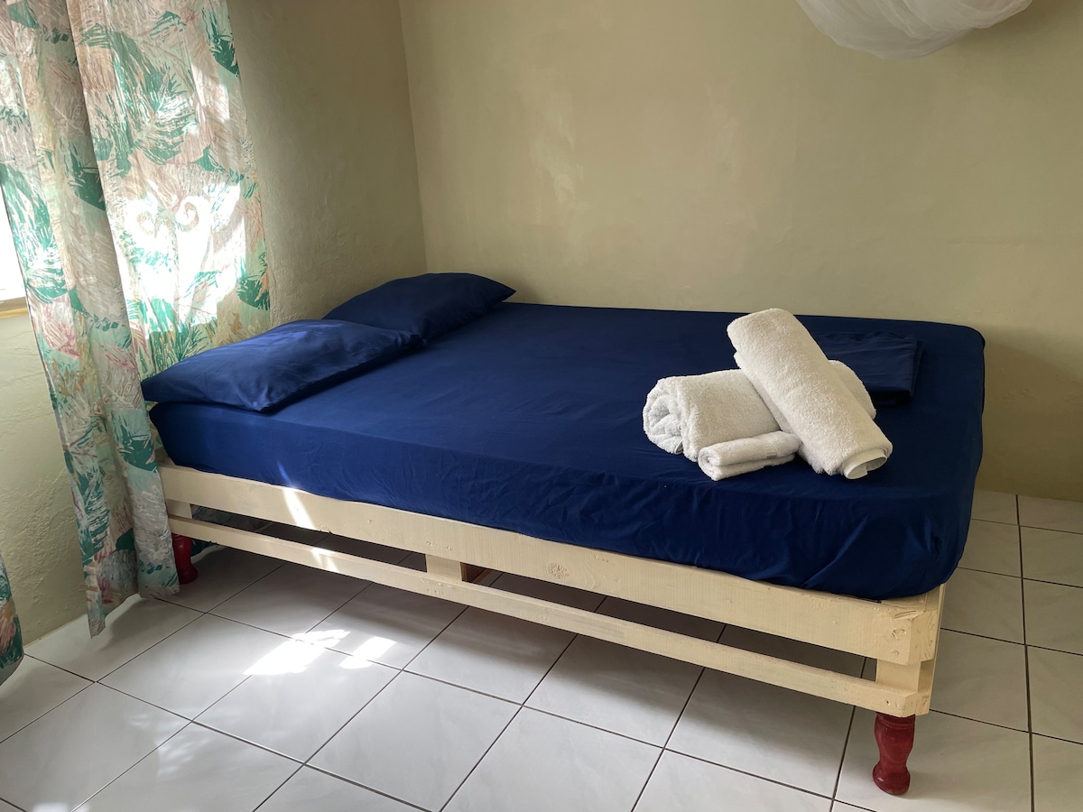 A simple sleeping area is furnished with a spacious bed dressed in a blue fitted sheet. Fresh white towels are neatly arranged on the bed. Natural light filters through a sheer curtain adorned with a tropical leaf design, illuminating the tiled floor.