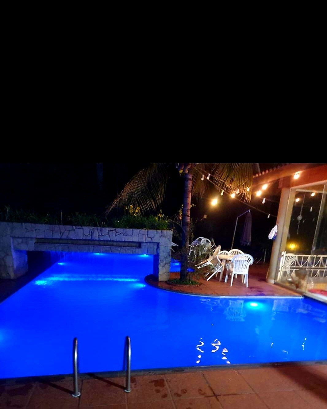 A tranquil pool area is illuminated in soft blue light, with steps leading into the water. A spacious deck features white chairs and an inviting atmosphere, surrounded by lush greenery and gentle lighting. The serene setting is ideal for evening relaxation.
