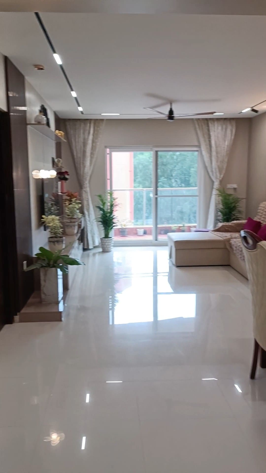 A bright living area features a comfortable sofa, adorned with cushions, inviting relaxation. Large glass doors lead to a balcony, allowing natural light to fill the space. Potted plants line the area, contributing to a fresh atmosphere, while elegant curtains frame the windows.