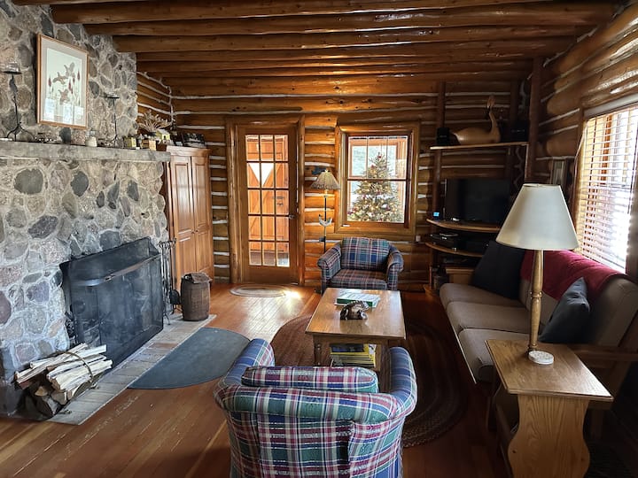 Main sitting area around stone fireplace. 3-season porch (Christmas tree during holiday season) in background, the front entrance to the cabin.