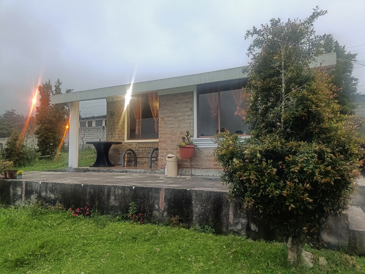 A charming cabin is set against a green landscape, featuring large windows that allow natural light to fill the interior. A table and chairs are positioned on a spacious patio, surrounded by well-maintained plants and trees. Soft lighting adds a welcoming glow to the exterior.