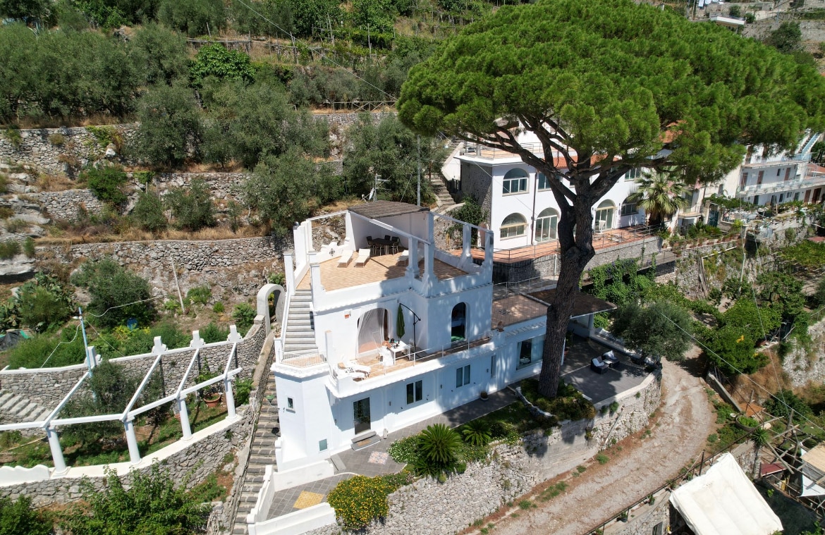 The villa is framed by a centuries-old pine tree, highlighting its distinct architectural features. The structure showcases multiple levels with open terraces, allowing for scenic views. Surrounding greenery complements the white facade, providing a serene Mediterranean atmosphere.