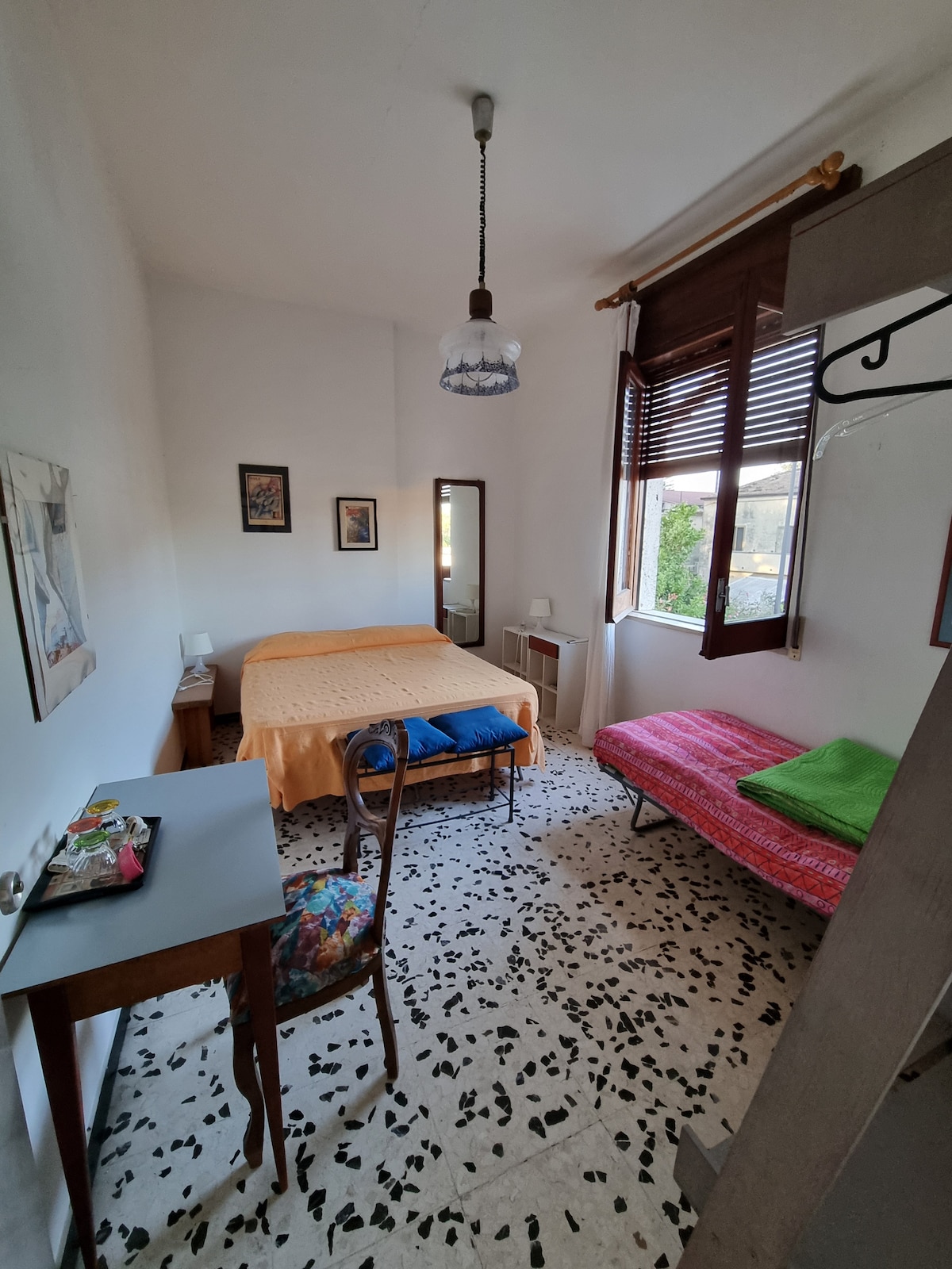 A spacious room features a light-colored wall and a large bed covered with a bright orange blanket. A small desk is situated next to a chair with a floral design. A single futon is placed to the side. Natural light enters through two windows adorned with wooden shutters.