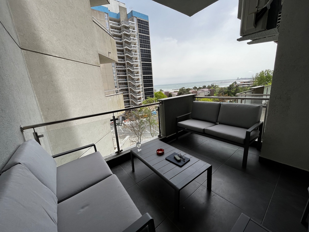 A spacious balcony is furnished with a modern seating arrangement that includes a coffee table and two sofas. The view overlooks a cityscape with visible buildings and the sea in the distance. A calming atmosphere is enhanced by the presence of greenery.