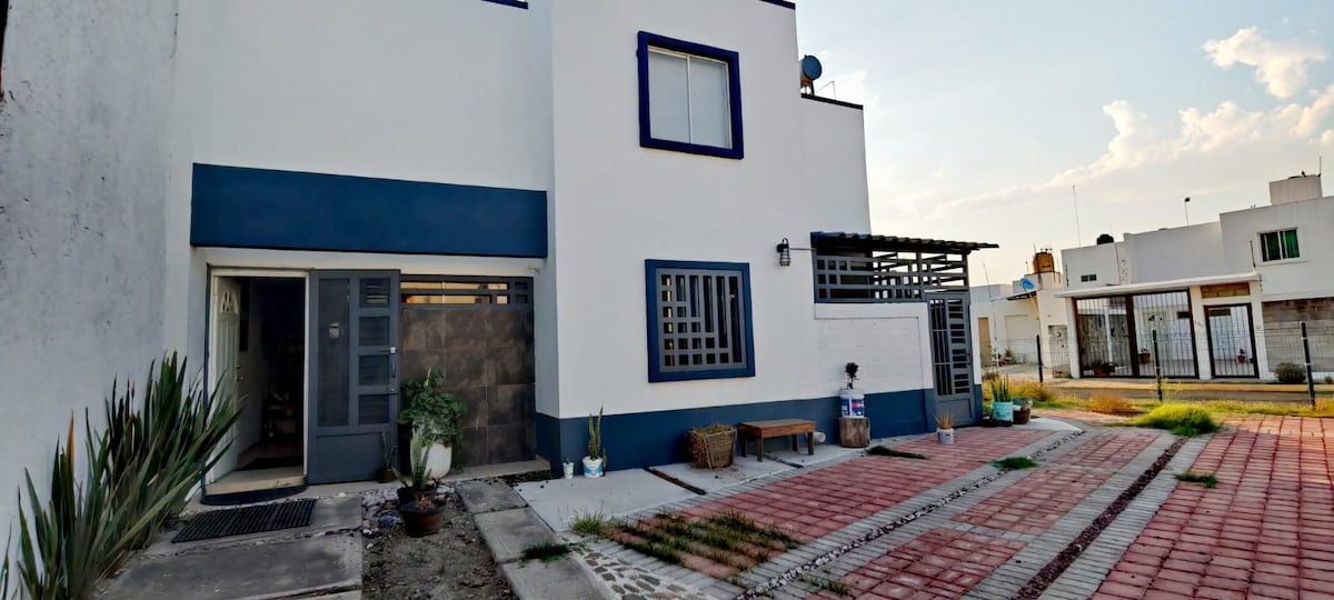 An exterior view of a modern two-story building is presented, featuring a light-colored facade with blue accents. A well-maintained pathway leads to a covered entrance area, adorned with potted plants. The surrounding outdoor space includes a tiled surface, enhancing the entryway's welcoming appearance.
