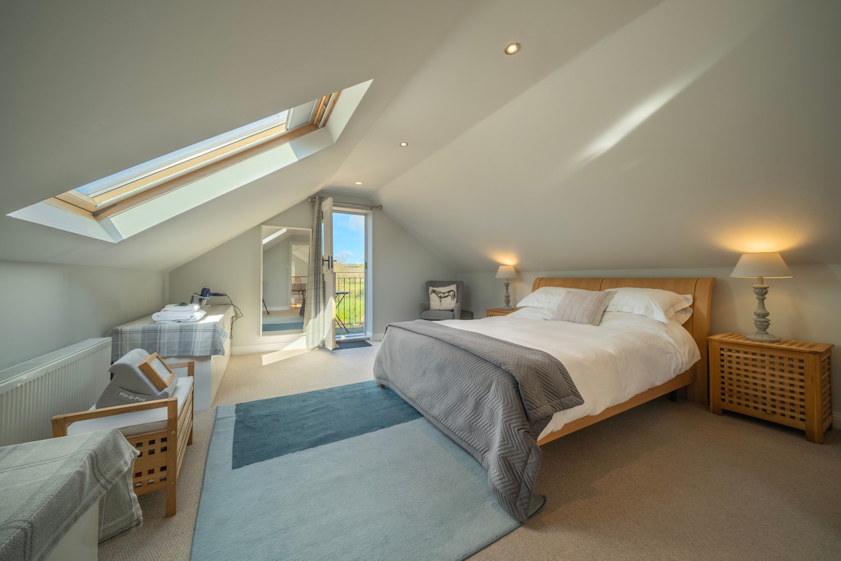 A well-lit bedroom features a spacious bed with crisp white linens and soft grey accents. A large skylight provides natural light, while a balcony door offers access to outdoor views. Two bedside lamps are positioned beside woven storage baskets, enhancing the room's serene atmosphere.