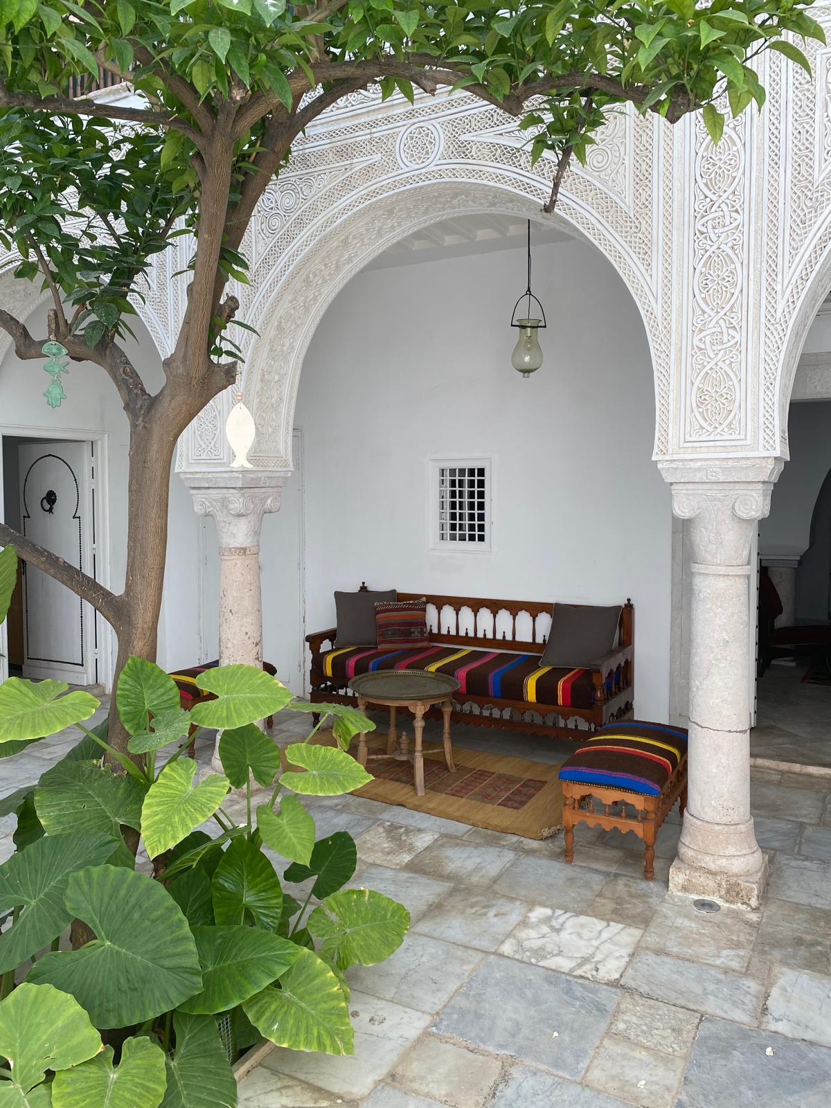 An inviting seating area is framed by an ornate archway, featuring a wooden bench adorned with colorful cushions. A small table sits between the bench and a vibrant striped stool. Lush green plants and a tree add a touch of nature to the space, complemented by traditional tiled flooring.