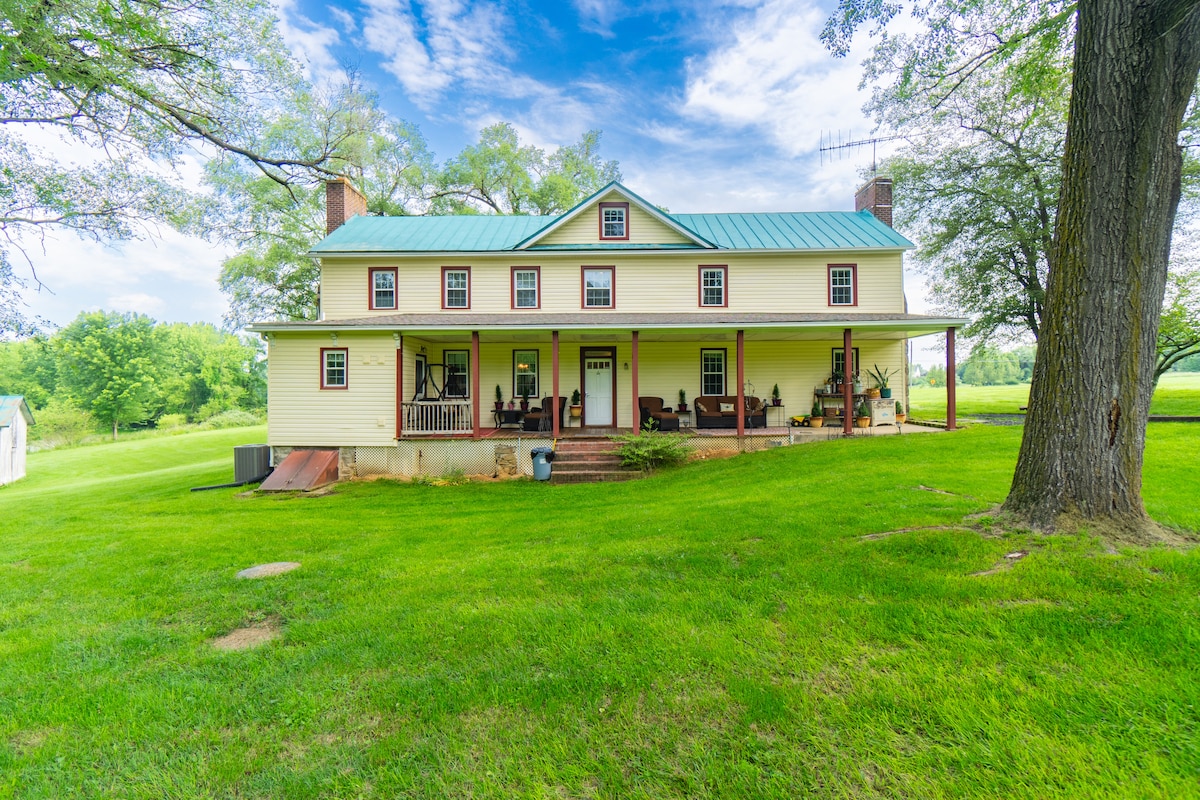 The yellow farmhouse, featuring a green metal roof, is set against a backdrop of lush greenery. A spacious covered porch extends across the front, adorned with chairs and a welcoming atmosphere, inviting guests to relax and enjoy the surrounding open spaces.