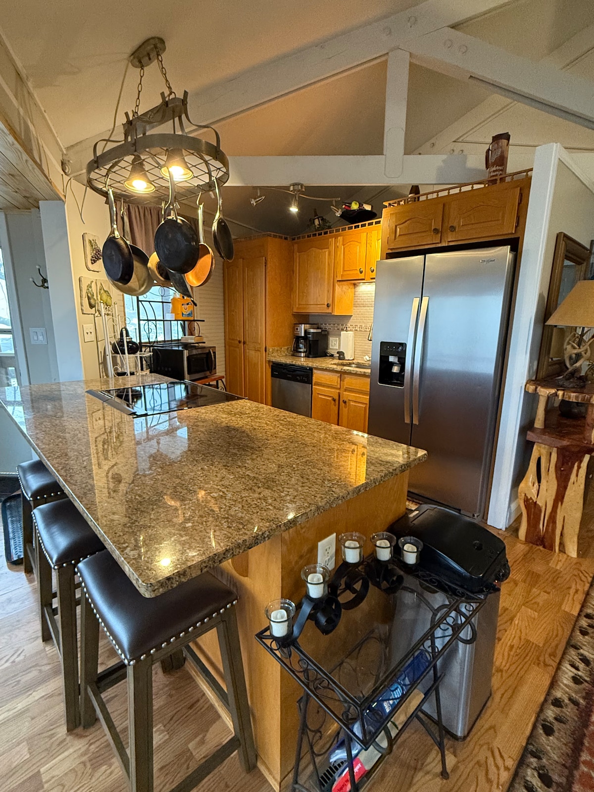 A fully-equipped kitchen showcases wooden cabinetry and modern stainless steel appliances. A spacious island with seating for four is complemented by hanging cookware. Natural light filters through a nearby window, illuminating the warm tones of the granite countertops and wooden floors.
