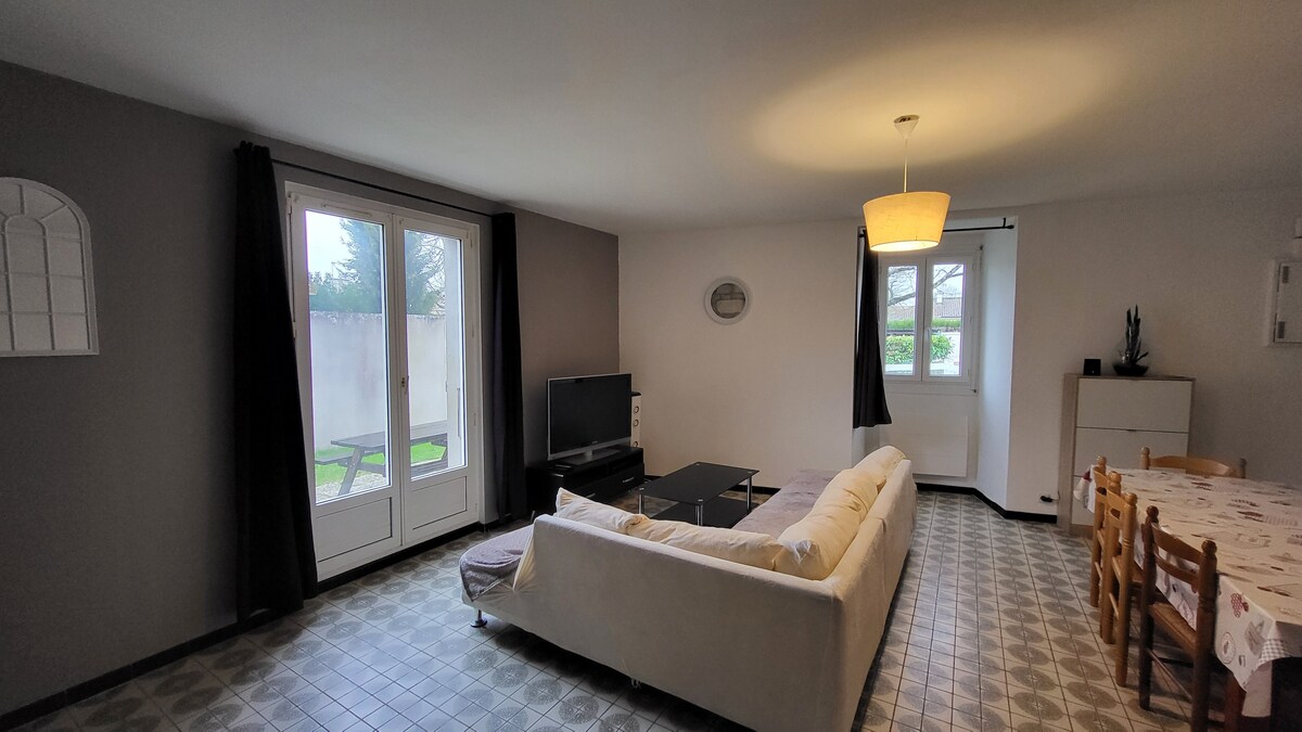 A light-filled living area is featured, with a spacious corner sofa facing a television. Large glass doors provide views of a private terrace and garden. A dining table with wooden chairs is positioned nearby, complementing the neutral color scheme of the room.