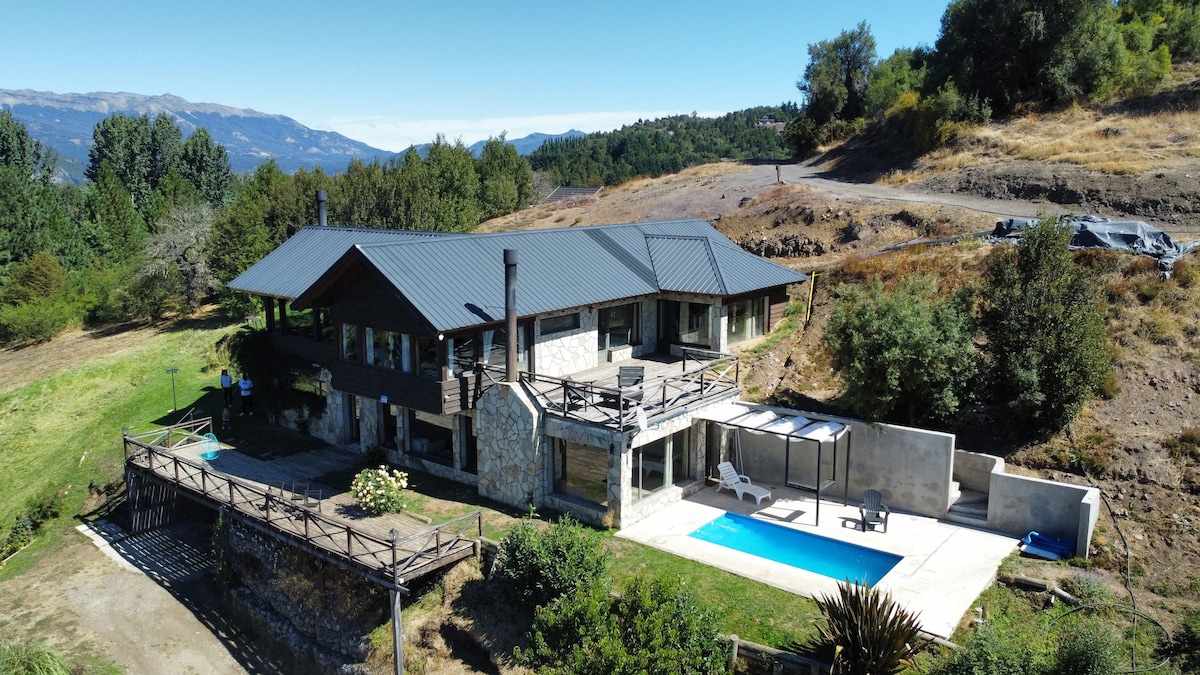 The two-story house features a combination of stone and wooden elements, set against a backdrop of mountains. A private pool area is visible, accompanied by lounge chairs, while landscaped greenery surrounds the property. The expansive deck showcases outdoor space, ideal for relaxation.
