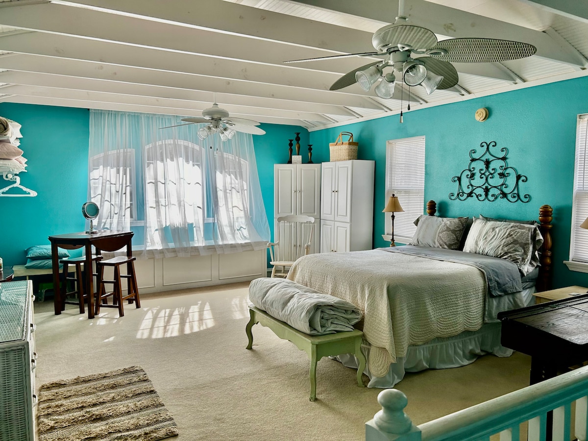 A spacious bedroom featuring a vaulted ceiling and bright turquoise walls. A large bed is positioned centrally, complemented by a side table with a lamp. A small dining table with chairs is visible, along with a dresser and ample natural light filtering through multiple windows.